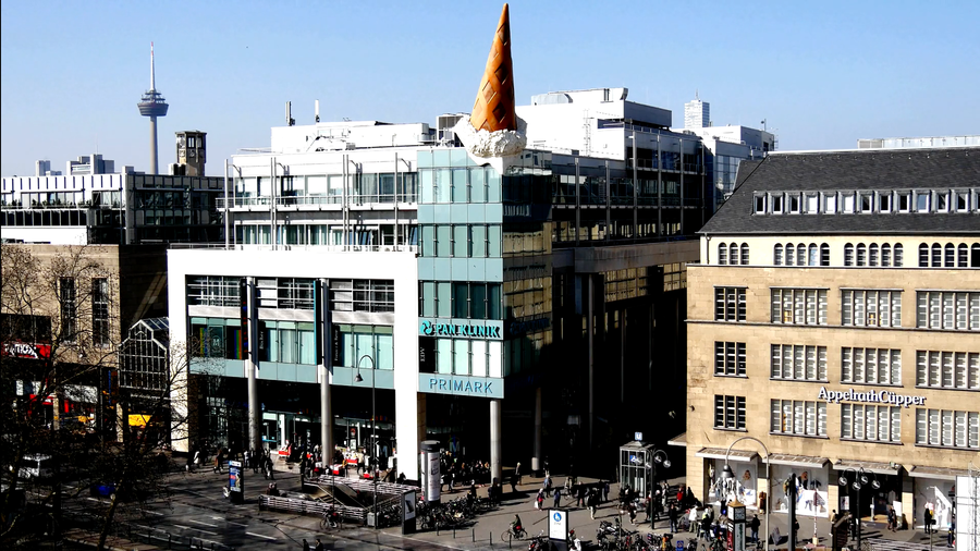 In der Stadtansicht ist ein modernes Gebäude mit einem großen, bunten Eisbecher-Dekor auf dem Dach zu sehen. Vor dem Gebäude sind Menschen zu sehen, die sich auf dem Platz bewegen, während im Hintergrund ein Aussichtsturm sichtbar ist.