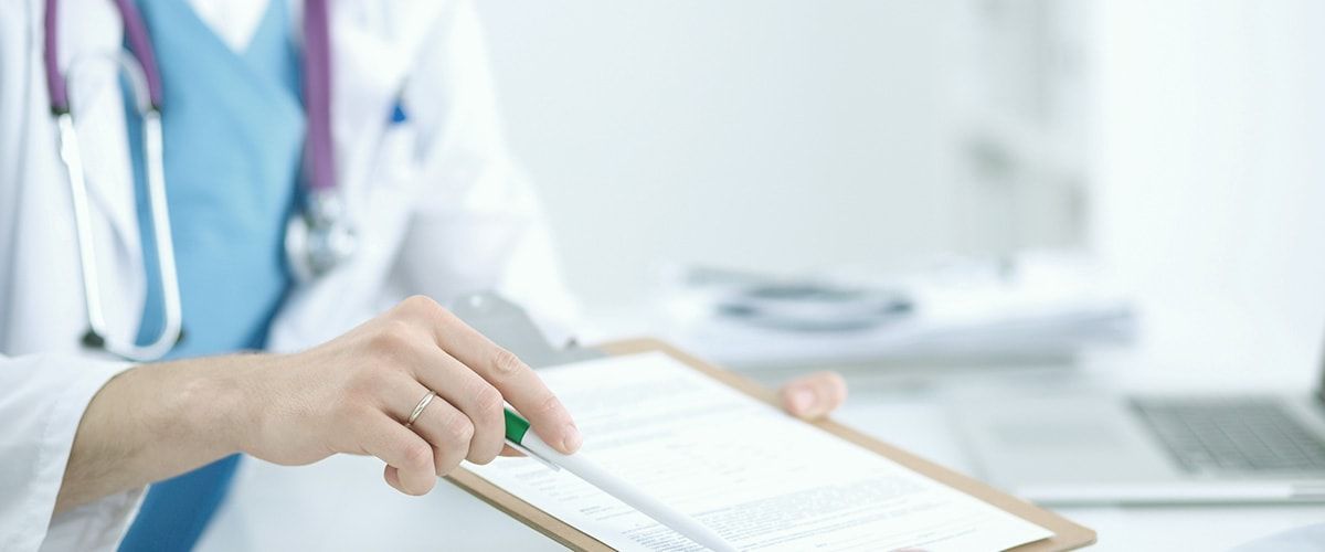 Ein Arzt in einem weißen Kittel hält ein Clipboard und zeigt mit einem Stift auf ein Dokument. Im Hintergrund sind verschwommene medizinische Utensilien und ein Laptop zu sehen. Das Bild vermittelt eine professionelle medizinische Umgebung.